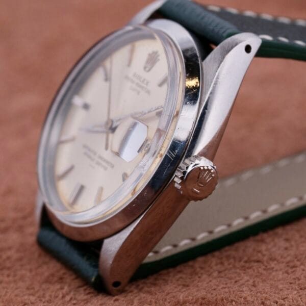 Close-up of a Rolex Oyster Perpetual Date 1500 Silver Dial wristwatch with a silver case and cream dial featuring a date display. Resting on a soft brown surface, the watch flaunts a green leather strap, while the crown showcases its distinctive emblem.