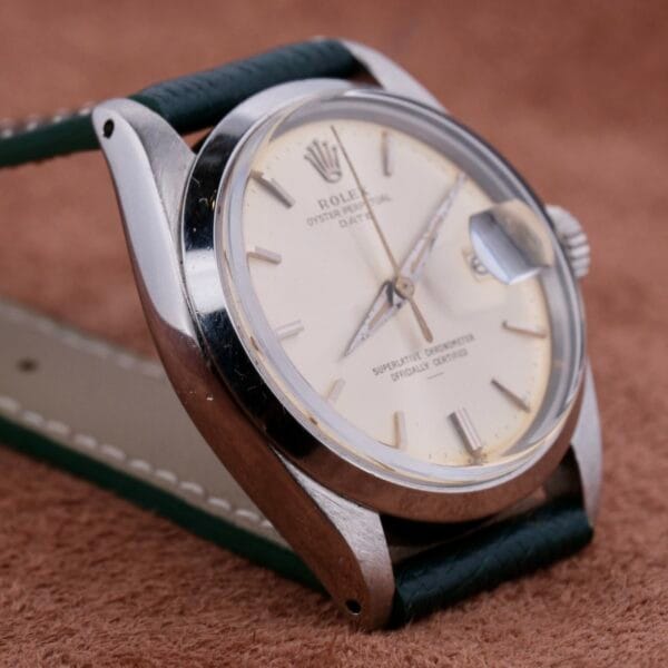 Close-up of a Rolex Oyster Perpetual Date 1500 with a silver case and cream dial featuring a date display at 3 oclock. It has a dark green leather strap and rests elegantly on a soft brown surface.