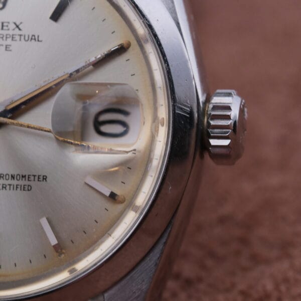 A close-up of the Rolex Oyster Perpetual Date 1500 Silver Dial reveals the brand label on its face. The time is around 10:08, and through its magnifying lens, the date window shows the number 6.