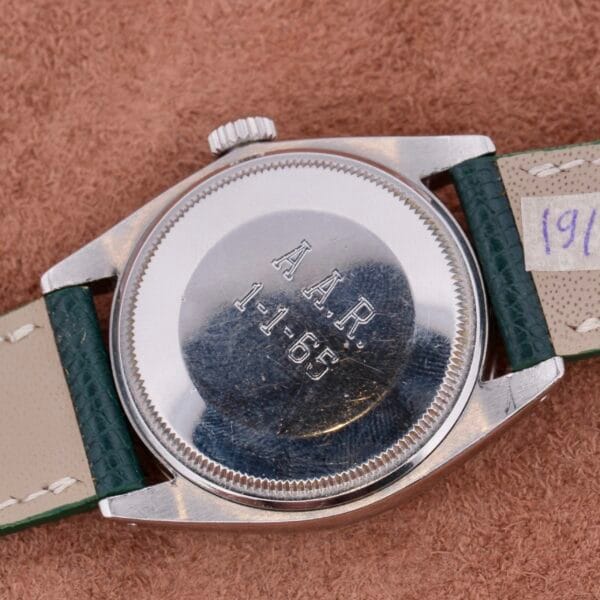On a tan surface rests a Rolex Oyster Perpetual Date 1500 Silver Dial, elegantly inscribed with A.A.R. 1-1-65. Its dark green leather strap hints at its legacy, with part of another marking intriguingly obscured, blending mystery and timeless style.