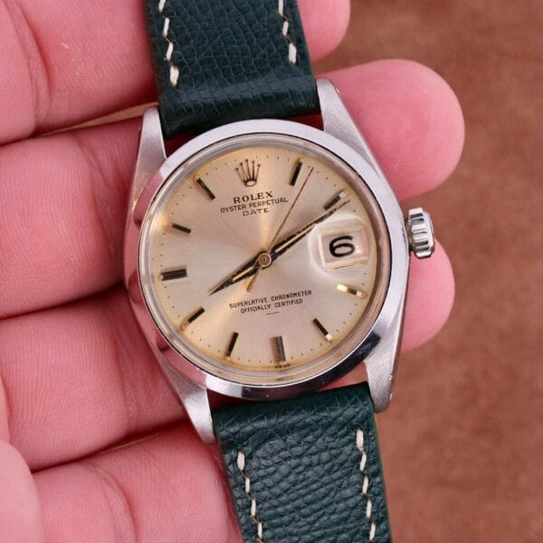 A hand elegantly holds a Rolex Oyster Perpetual Date 1500 Silver Dial watch with a green leather strap, silver case, and beige dial. Roman numerals and clock hands grace the face, while the date display shows the number 6.