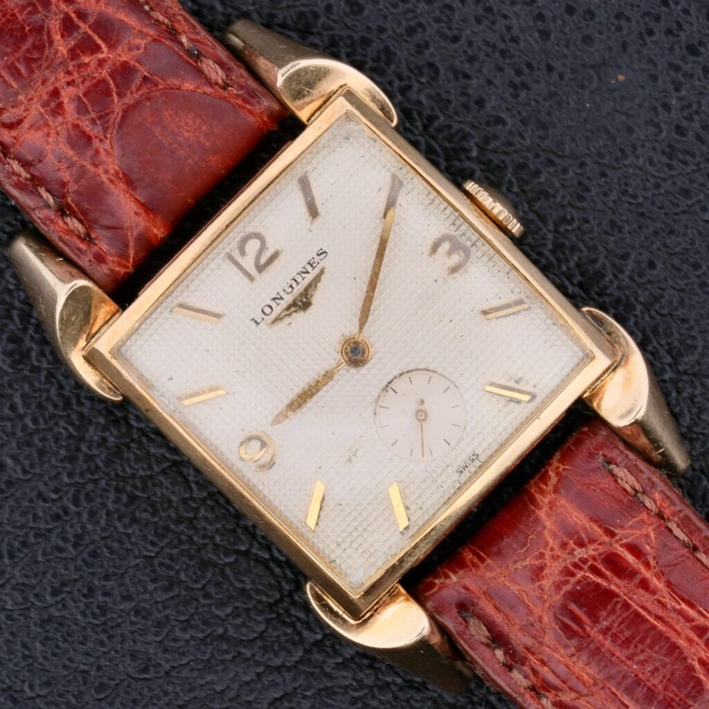 A Longines Franklin Square Waffle Dial Cal 23Z wristwatch with a gold case and brown leather strap rests on a dark textured surface.