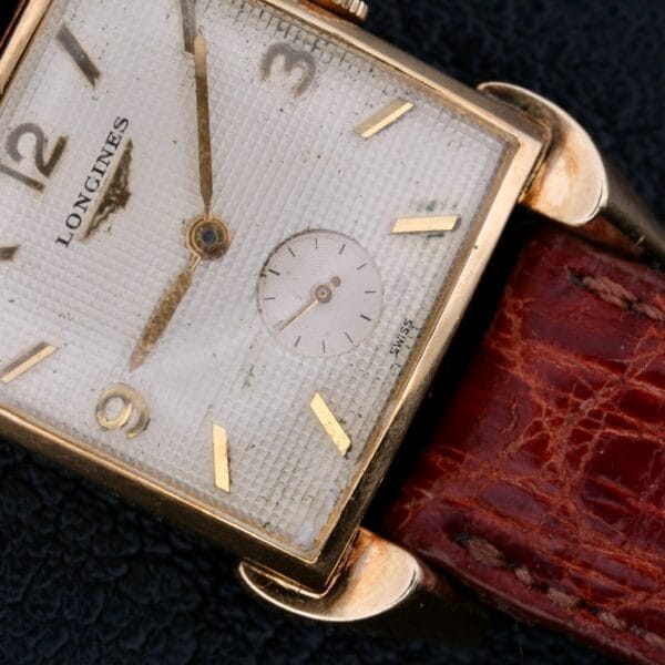 Close-up of a Longines Franklin Square Waffle Dial Cal 23Z watch, featuring a square, gold-tone case and textured white dial. Gold numerals complement the sub-dial, while the worn brown leather strap adds charm. The hands display approximately 8:57.