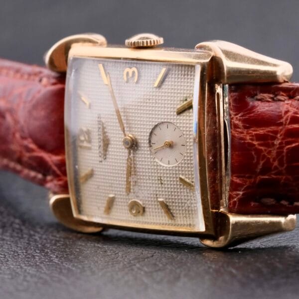 A close-up of the Longines Franklin Square Waffle Dial Cal 23Z wristwatch showcases its vintage rectangular design with a gold casing. It features a textured dial with gold markers and hands, attached to a worn brown leather strap against a dark background.