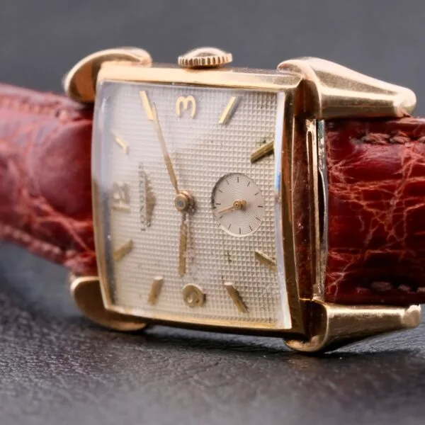 A close-up of the Longines Franklin Square Waffle Dial Cal 23Z wristwatch showcases its vintage rectangular design with a gold casing. It features a textured dial with gold markers and hands, attached to a worn brown leather strap against a dark background.