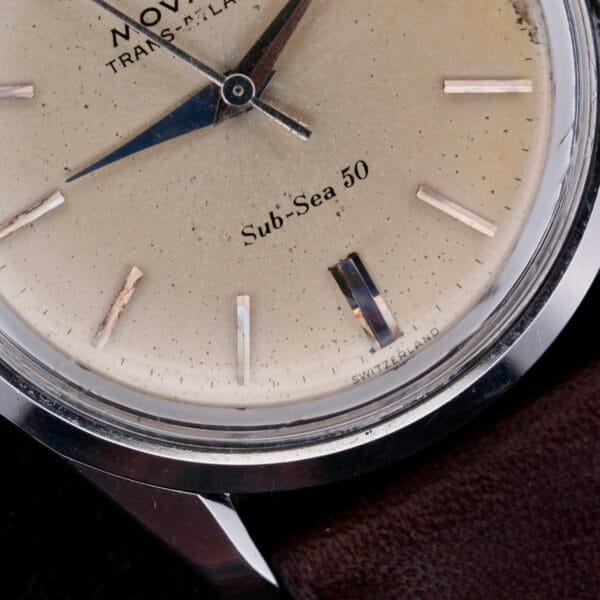 Close-up of the vintage Movado Trans-Atlantic Sub Sea 50 watch with an off-white dial, dark brown leather strap, silver hour markers, and a small second hand. The scratched glass reveals text reading Sub-Sea 50 and Switzerland.