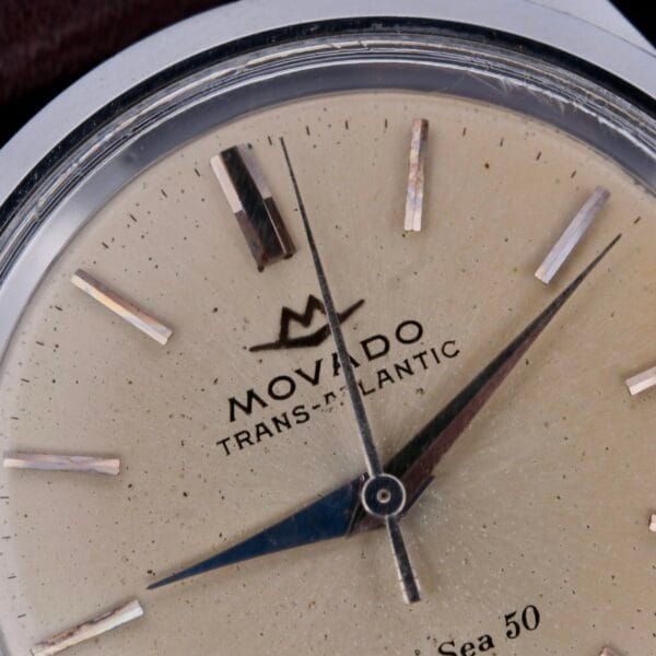 Close-up of a Movado Trans-Atlantic Sub Sea 50 watch face, featuring silver indices and hands on a beige dial with Movado Trans-Atlantic and Sea 50 text. The casing has a silver tone.