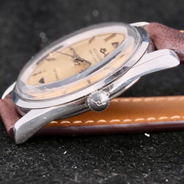 The Movado Trans-Atlantic Sub Sea 50 watch showcases a tan dial with triangular hour markers, encased in sleek silver and paired with a brown leather strap against a speckled dark background, enhancing its timeless appeal.