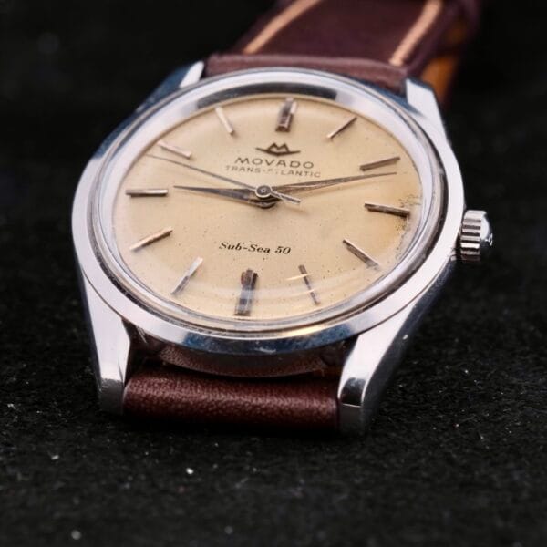 Close-up shot of the Movado Trans-Atlantic Sub Sea 50 watch, featuring a beige dial, silver indices, and a brown leather strap, against a black background.