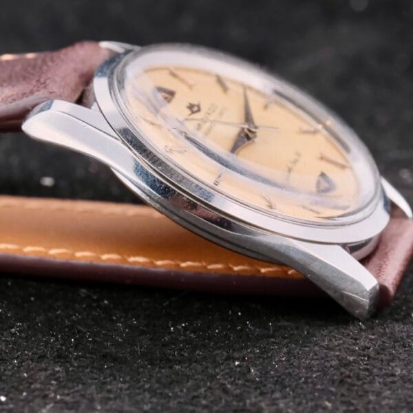 Close-up of a Movado Trans-Atlantic Sub Sea 50 vintage wristwatch, featuring a round beige dial, silver casing, and a brown leather strap with visible stitching. Its set against a textured dark surface, showcasing numbers and markers for precise timekeeping.