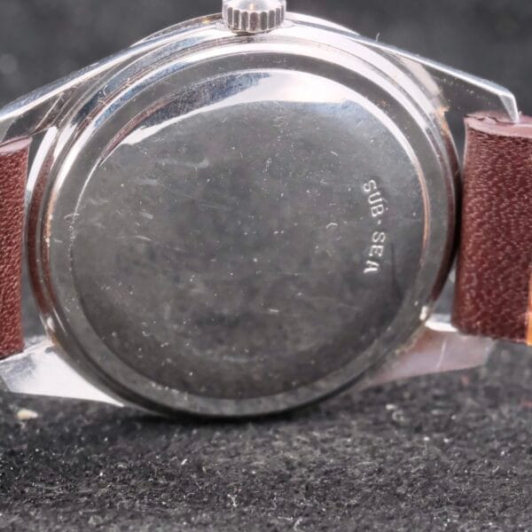 A close-up highlights the engraved SUB-SEA on the back of a polished metal Movado Trans-Atlantic Sub Sea 50 wristwatch, featuring a brown leather strap. It rests gracefully on a dark textured surface.
