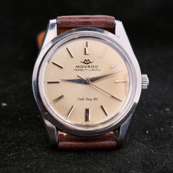 A vintage Movado Trans-Atlantic Sub Sea 50 watch, featuring a cream dial, silver hour markers, and a brown leather strap, stands elegantly against a dark background.