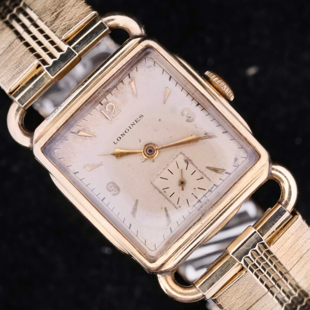The Longines Square Driver Lugs Cal 10L is a vintage wristwatch featuring a square gold case, beige dial, gold hands, Arabic numerals at 12, 3, 6, and 9 with a sub-dial above 6. It comes with an elegant textured gold-tone bracelet.