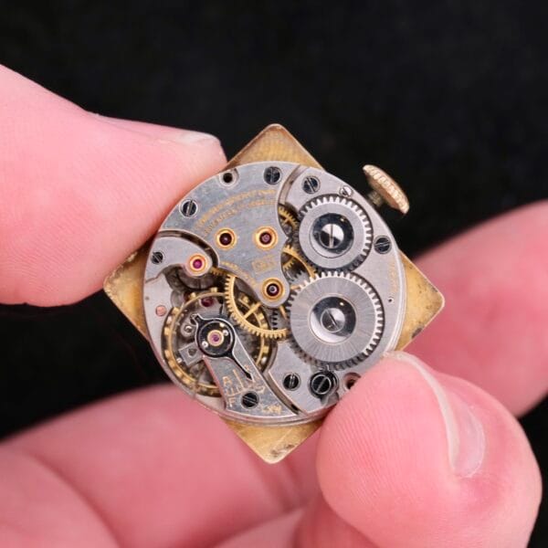 A close-up shows a persons hand holding the intricate watch movement of a Longines Square Driver Lugs Cal 10L, with gears and mechanisms visible against a dark background.