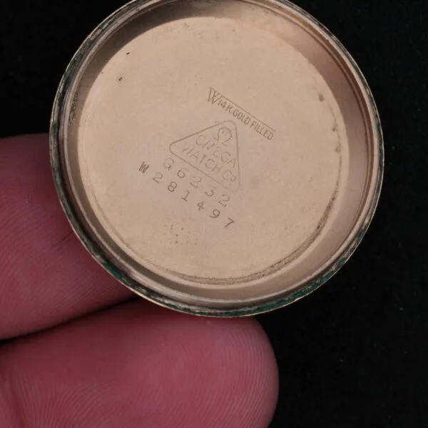 A close-up shows a hand holding the inside of an Omega Automatic Bumper watch case, marked 14K Gold Filled, with the Omega Watch Co triangle logo and serial numbers G6232 and W281497 against a black background.