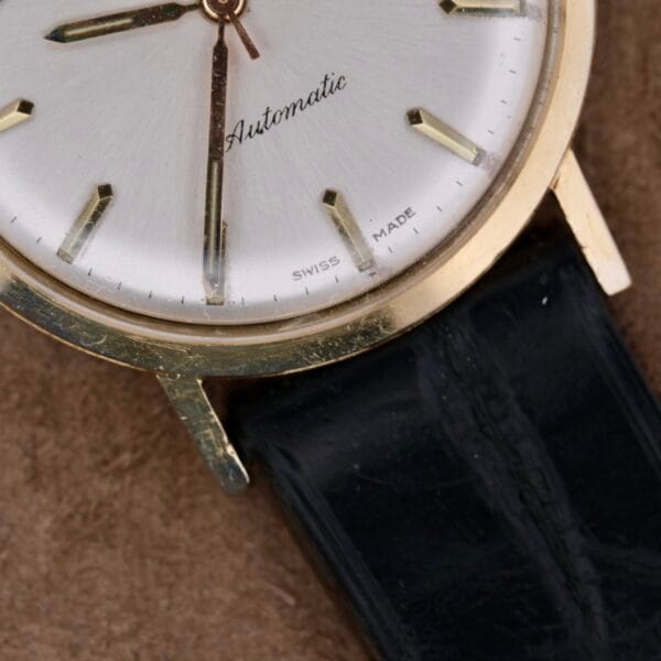 A close-up of the vintage Zodiac Hermetic 14k Gold Automatic Date wristwatch features a gold-tone case, black leather strap, silver face with gold hour markers and hands. Automatic is at the center and Swiss Made at the bottom.
