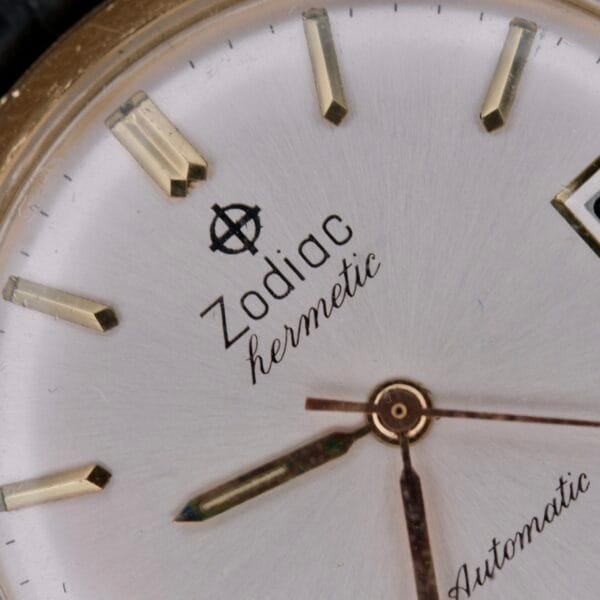 Close-up of a Zodiac Hermetic 14k Gold watch face with gold hour markers and hands, displaying Zodiac, hermetic, and Automatic. A portion of the date window appears on the right.