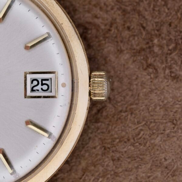 Close-up of a Zodiac Hermetic 14k Gold Automatic Date wristwatch on a brown textured background. It features a silver dial displaying the date as the 25th, with gold hour markers and hands enhancing its classic design.