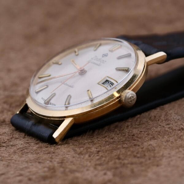 A Zodiac Hermetic 14k Gold Automatic Date wristwatch, featuring a gold-tone case, black leather strap, thin hour markers, date display, and two hands, rests on a textured brown surface.