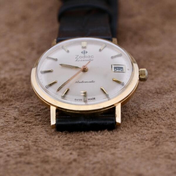 The Zodiac Hermetic 14k Gold Automatic Date, a vintage wristwatch with a 14k gold-tone case and black leather strap, is showcased on a textured brown surface. It features a white dial with gold markers, gold hands, and a date window at the 3 oclock position.