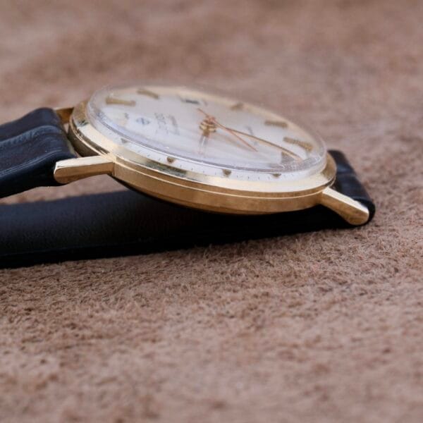 A close-up side view of a Zodiac Hermetic 14k Gold Automatic Date wristwatch with a black leather strap on a textured brown surface, featuring gold hour markers and hands.