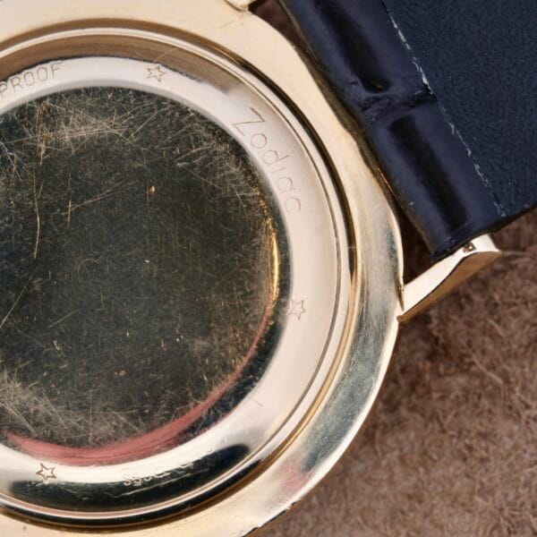 A close-up of the Zodiac Hermetic 14k Gold Automatic Date wristwatch reveals its 14k gold casing with visible scratches and engraved Zodiac among other inscriptions. The watch boasts a sleek black leather strap.