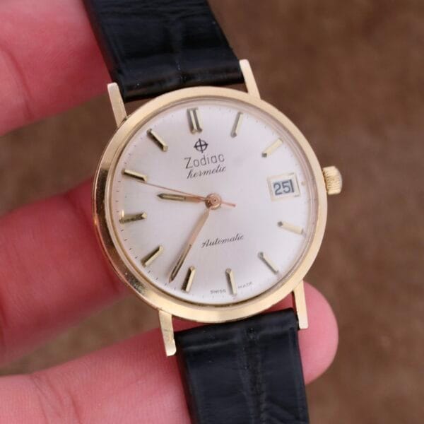 Close-up of the Zodiac Hermetic 14k Gold Automatic Date wristwatch with a white dial, black leather strap, gold hour markers, and a date window showing 25.