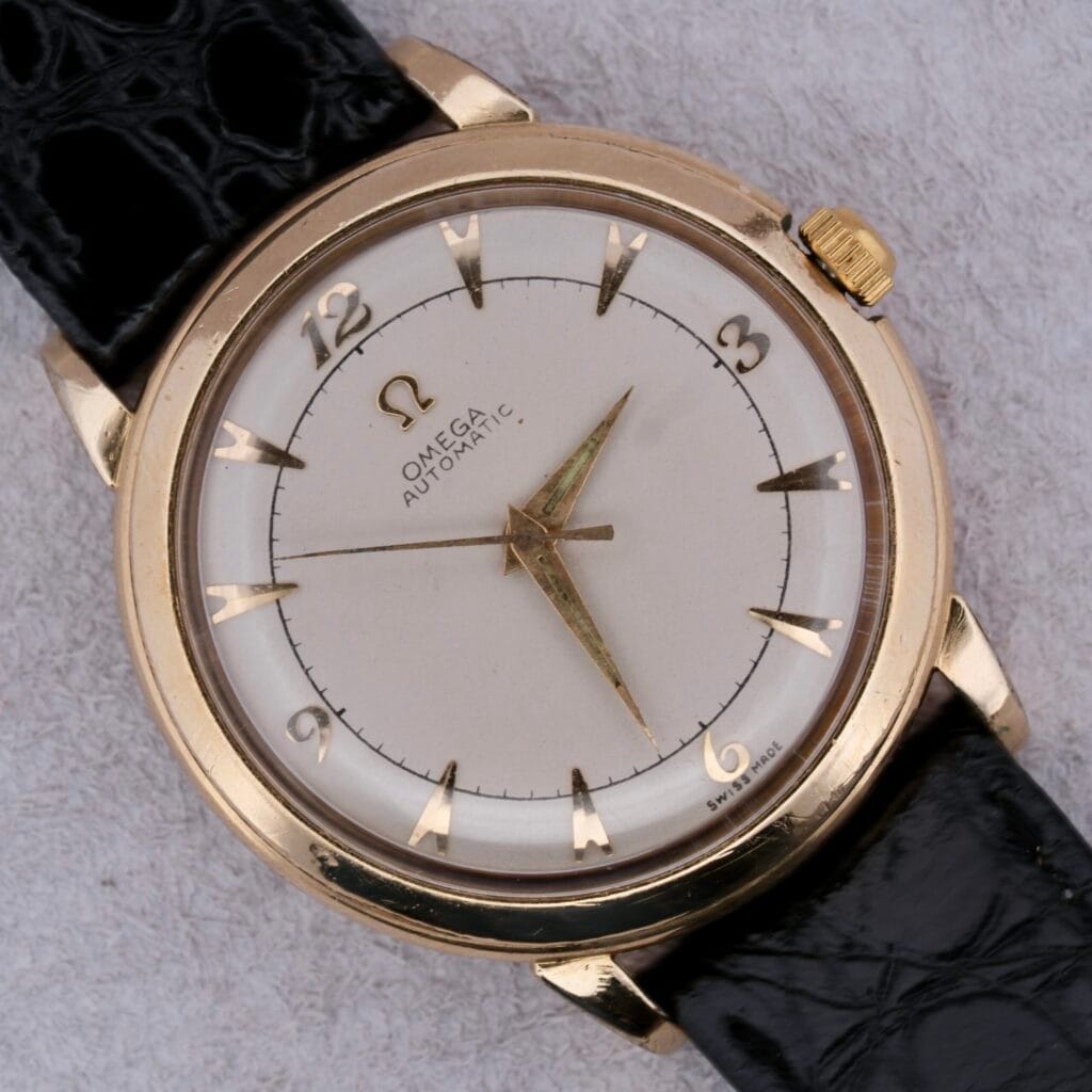 A vintage Omega Automatic Bumper watch boasts a round gold case and a white dial with gold hour markers, hands, and crown. It sits on a black leather strap with a textured light gray background.