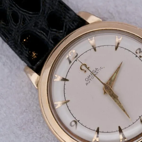 Close-up of an Omega Automatic Bumper watch featuring a gold-tone case, white dial, and black leather strap. It has thin gold hands, triangular hour markers, and the Omega logo prominently displayed on the dial.
