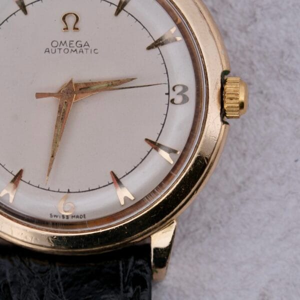 A close-up of the Omega Automatic Bumper wristwatch highlights its vintage charm with a gold case, white dial, black leather strap, and Swiss-made label. Gold hands and markers accentuate the Omega logo at 12 oclock.