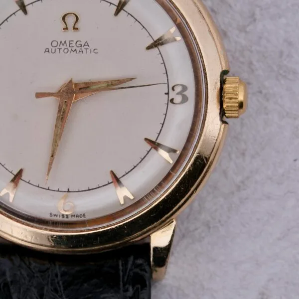 A close-up of the Omega Automatic Bumper wristwatch highlights its vintage charm with a gold case, white dial, black leather strap, and Swiss-made label. Gold hands and markers accentuate the Omega logo at 12 oclock.