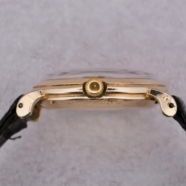 Side view of an Omega Automatic Bumper featuring a gold case and black leather strap. The side profile accentuates the crown and sleek design against a textured gray backdrop.