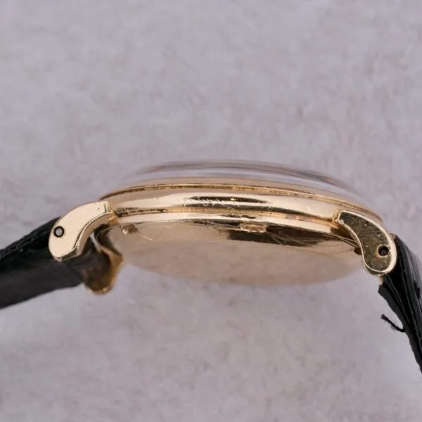 Side view of the Omega Automatic Bumper with a gold finish and thin black leather strap, featuring an elegant smooth, rounded casing against a textured gray background.