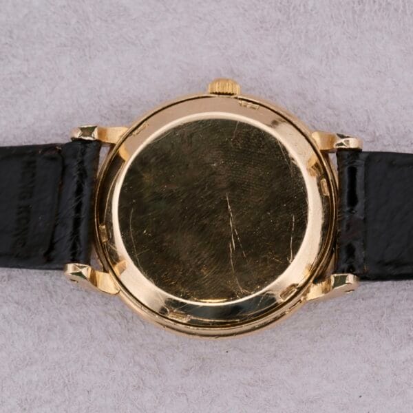 An Omega Automatic Bumper, featuring a scratched back casing and black leather strap, rests on a soft gray surface, with its round face not visible.