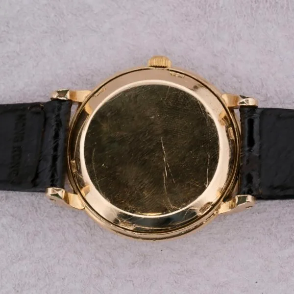 An Omega Automatic Bumper, featuring a scratched back casing and black leather strap, rests on a soft gray surface, with its round face not visible.