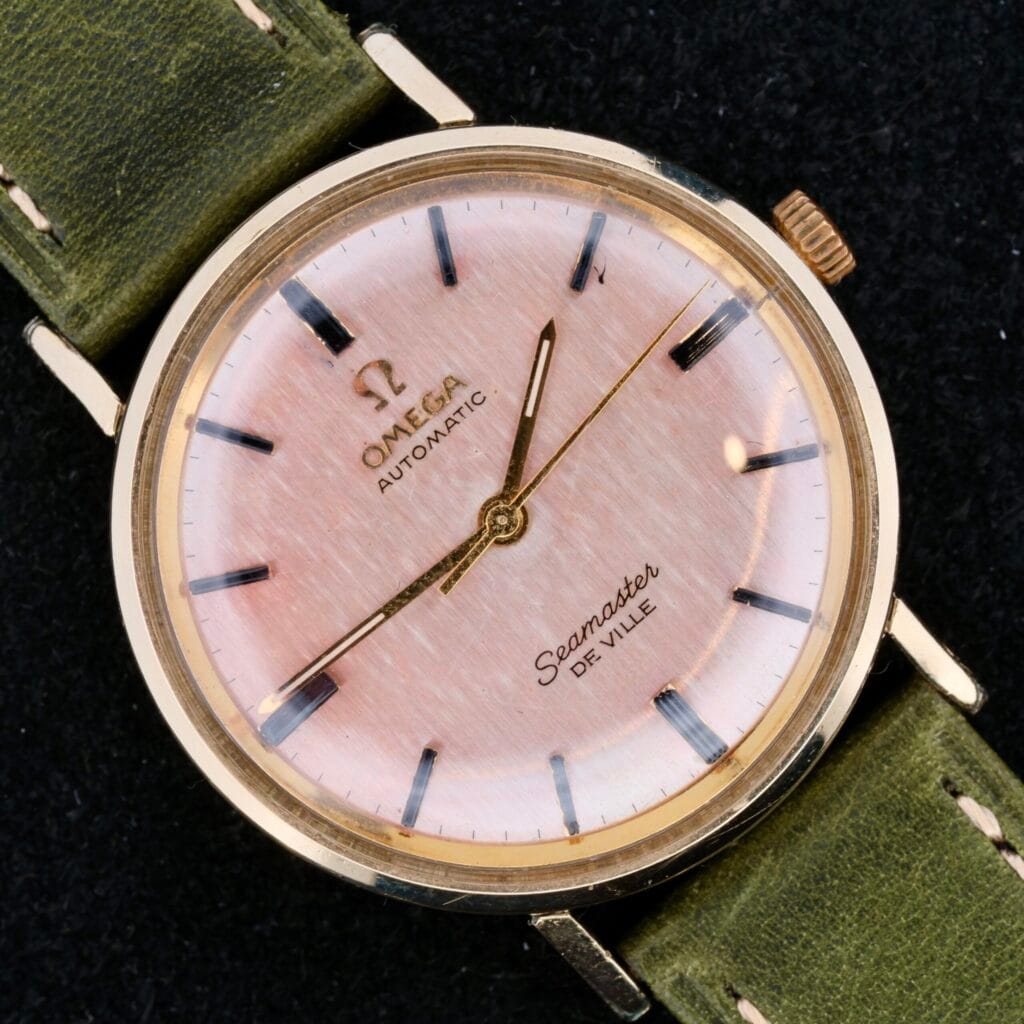 The Omega Seamaster Deville Automatic features a rose gold dial, gold casing, and gold hour markers. It includes a green leather strap and is showcased against a dark background.