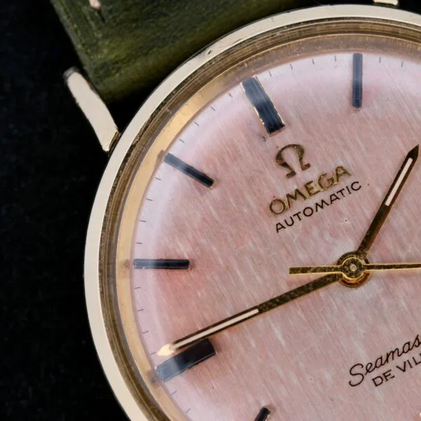 Close-up of an Omega Seamaster Deville Automatic Rose Gold Dial watch with a gold-tone case, rose dial, black hour markers, and Omega Automatic and Seamaster Deville branding. It features an elegantly dark green strap.