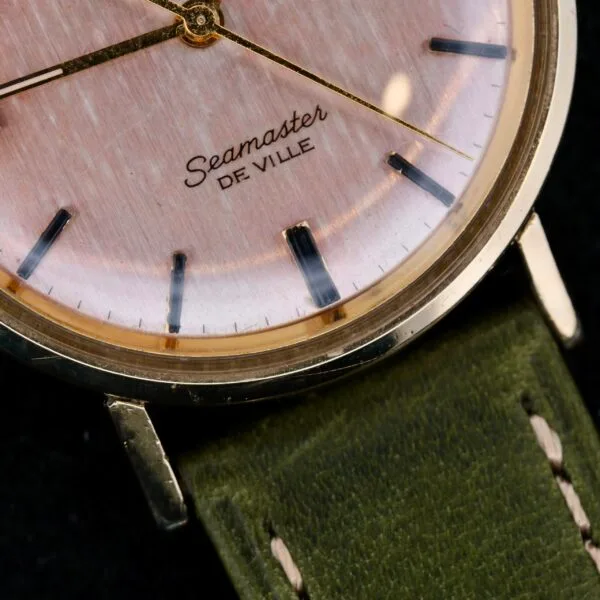 Close-up of an Omega Seamaster Deville Automatic Rose Gold Dial wristwatch, highlighting the Seamaster De Ville text. This watch boasts gold-tone hands, minimalist hour markers, and a gold-tone case elegantly matched with a green leather strap.