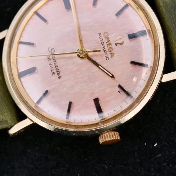 A close-up of the Omega Seamaster Deville Automatic Rose Gold Dial displays its round pinkish dial, gold-tone case, black hour markers, and gold hands, elegantly paired with a green leather strap against a dark background.