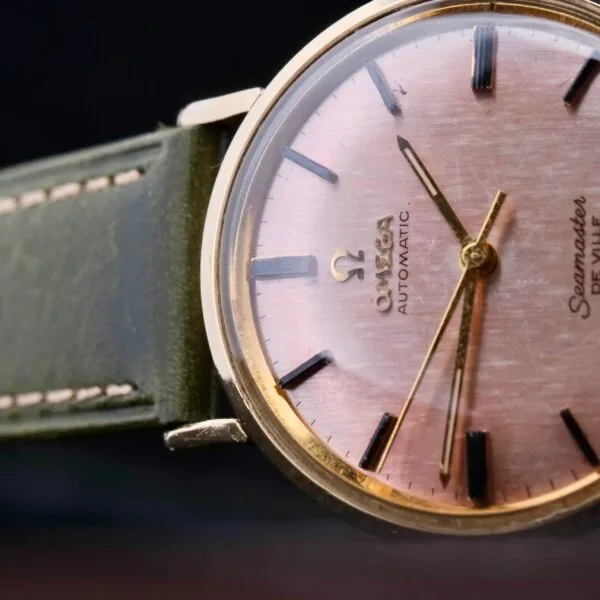 A close-up of an Omega Seamaster Deville Automatic features a rose gold dial with dark markers, gold casing, and hands. Showcasing Omega Automatic and Seamaster text, its paired with a green leather strap.