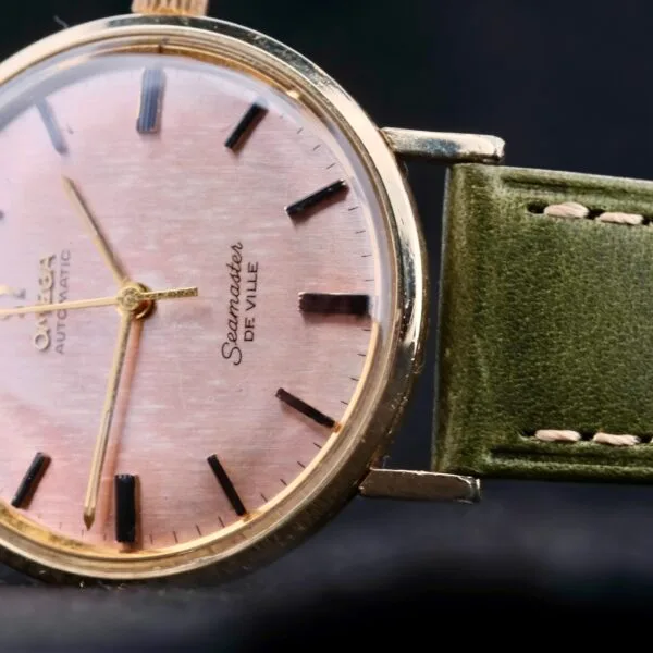 Close-up of an Omega Seamaster Deville Automatic with a rose gold dial, showcasing black hour markers and hands. The watch is encased in gold and features Automatic markings. A green leather strap with white stitching completes this timeless piece.