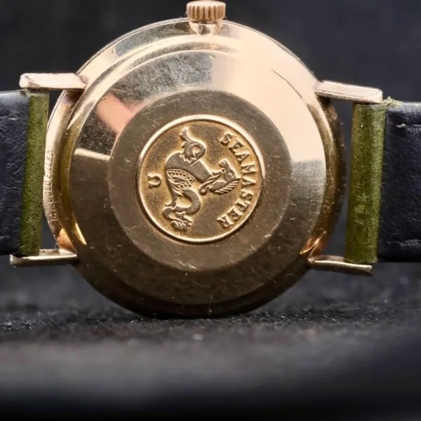 Close-up of the Omega Seamaster Deville Automatic Rose Gold Dial watch back, displaying a gold emblem with a seahorse and waves encircled by Seamaster. It has a dark leather strap with green lining, set against a black background.