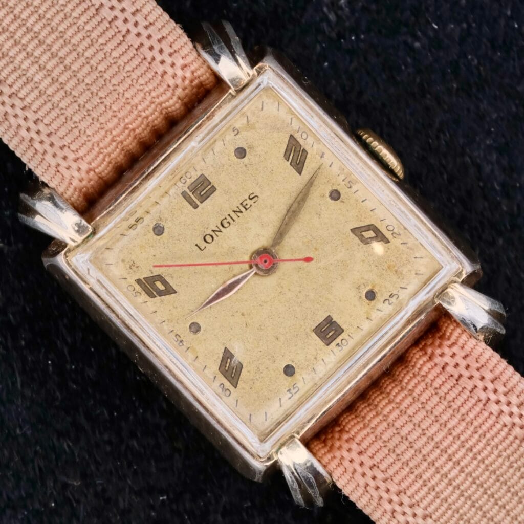 A Longines Square Gilt Dial Central Red Second wristwatch with a beige face and pink fabric strap, shown against a black background.