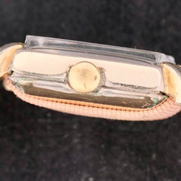 Side view of the Longines Square Gilt Dial Central Red Second, showcasing a vintage wristwatch with a rounded crown, gold-tone casing, and beige woven strap. The design features an acrylic crystal thats slightly raised against the dark, textured background.