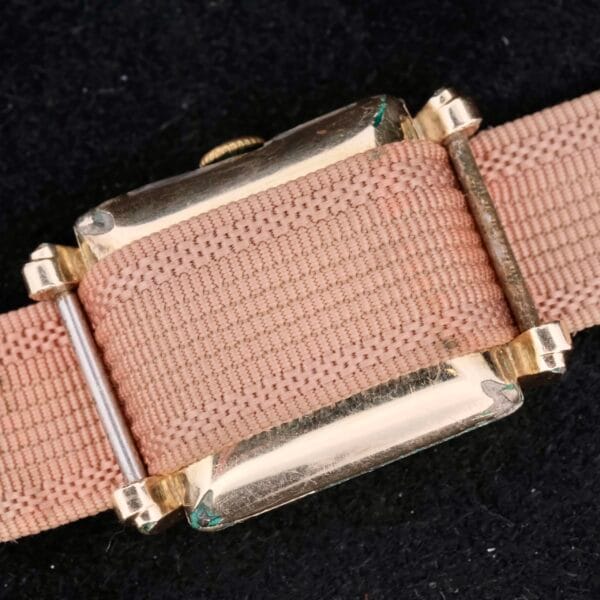 A close-up features a Longines Square Gilt Dial Central Red Second watch with a vintage gold case on a textured beige fabric strap, set against a dark, coarse background creating striking contrast with the rich gold and subtle beige tones.