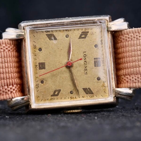 Close-up of a Longines Square Gilt Dial Central Red Second wristwatch with Arabic numerals and a beige fabric strap, set against a dark background. Signs of wear enhance its antique character.