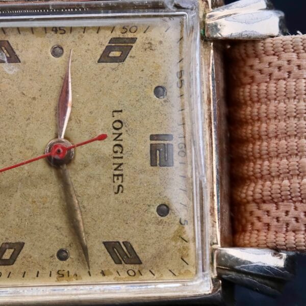 Close-up of a Longines Square Gilt Dial Central Red Second watch showcasing a square gilt dial with a textured beige face, red second hand, and black and gold hour markers. It features a tan fabric strap, and the case gracefully shows signs of aging.