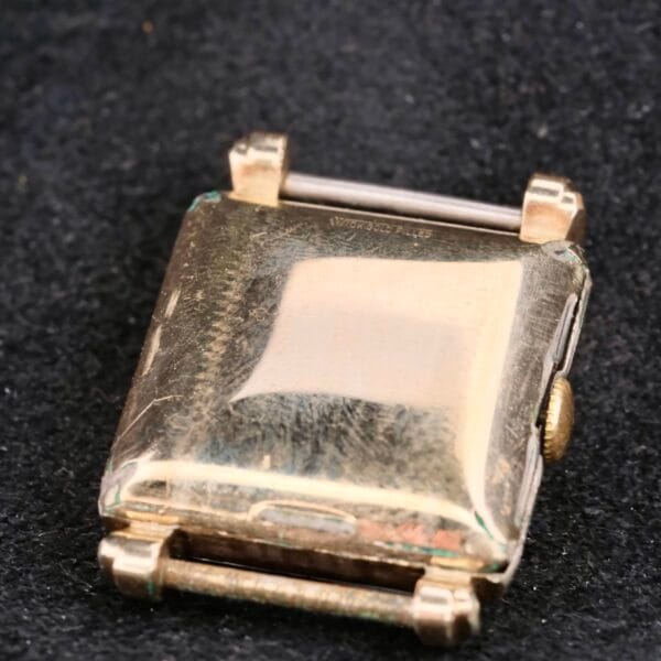 A vintage Longines Square Gilt Dial Central Red Second watch case rests on a dark, textured surface. Its rectangular shape is enhanced by a small winding crown on the right and visible top and bottom hinges, while the gold-toned metals wear and age evoke timeless elegance.