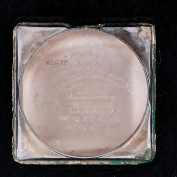 A scratched glass watch back with engraved text and numbers, partially legible. The aged and worn Longines Square Gilt Dial Central Red Second casing has a metallic frame. Its enigmatic presence against a black background hints at the history behind its gilt dial.