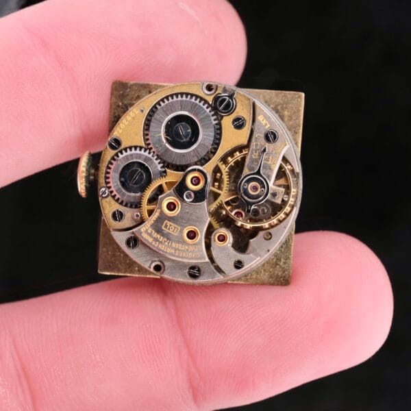 A close-up of fingers holding a Longines Square Gilt Dial Central Red Second mechanical watch movement displays the intricate gears and components, highlighting the craftsmanship of its inner workings.